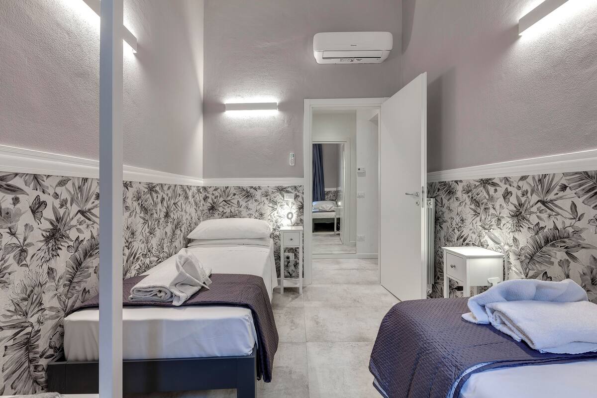 Une chambre d’hôtel avec deux lits, un papier peint à motifs floraux, une table de chevet avec une lampe et un appareil de climatisation fixé au mur.