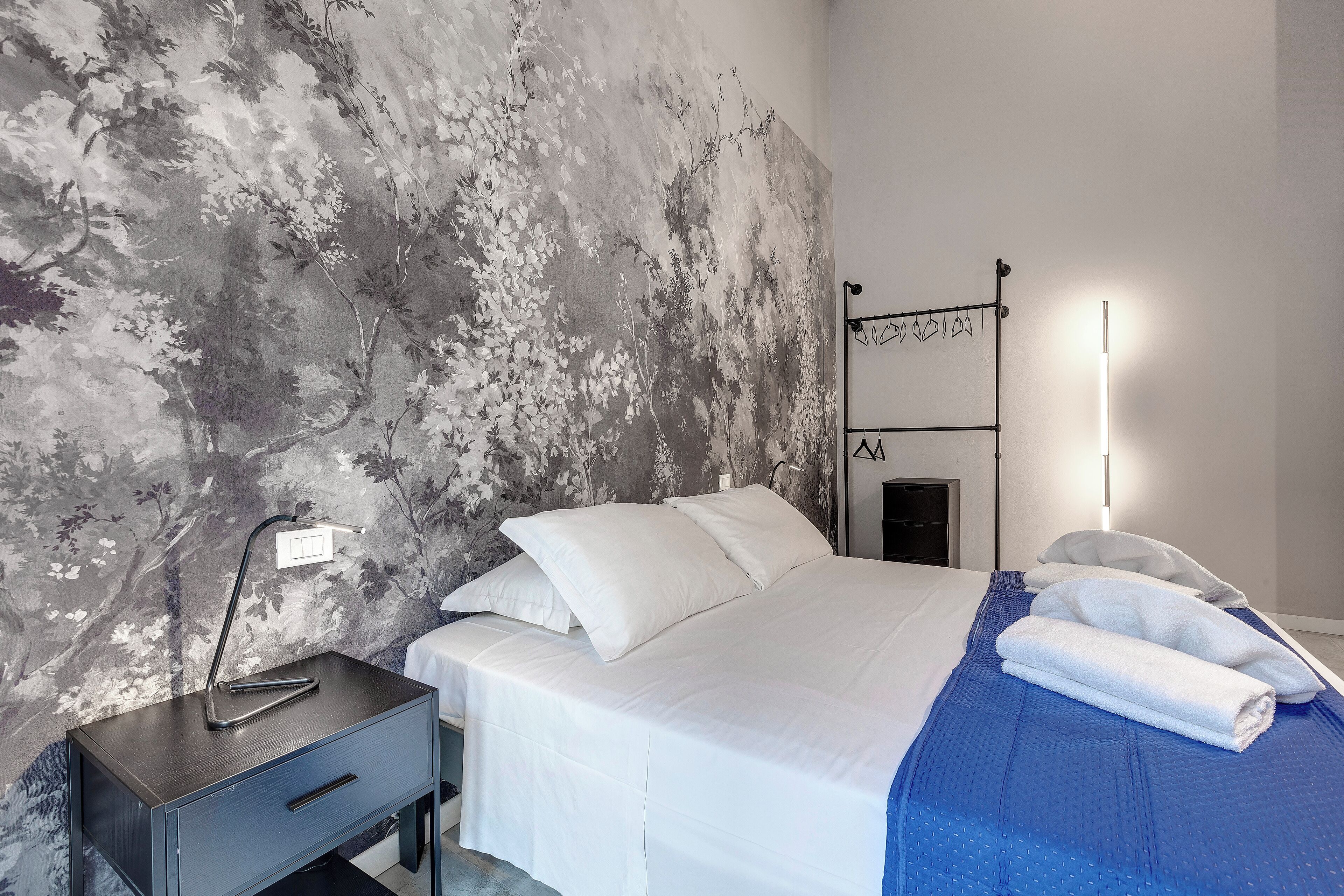 Un lit bien fait, avec du linge de lit blanc et une couverture bleue, une table de chevet avec une lampe, et un mur orné d’un motif floral.