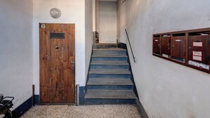 Un corridoio stretto con una porta in legno, un rilevatore di fumo metallico e una fila di armadietti in legno sulla parete destra.