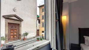 Une chambre d’hôtel avec un lit, une fenêtre avec des rideaux, une lampe et une vue sur la rue et les bâtiments environnants.