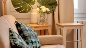 Ein beiges Sofa mit gemusterten Kissen, ein kleiner Holztisch mit Blumenvase und eine große grüne Pflanze im Hintergrund.