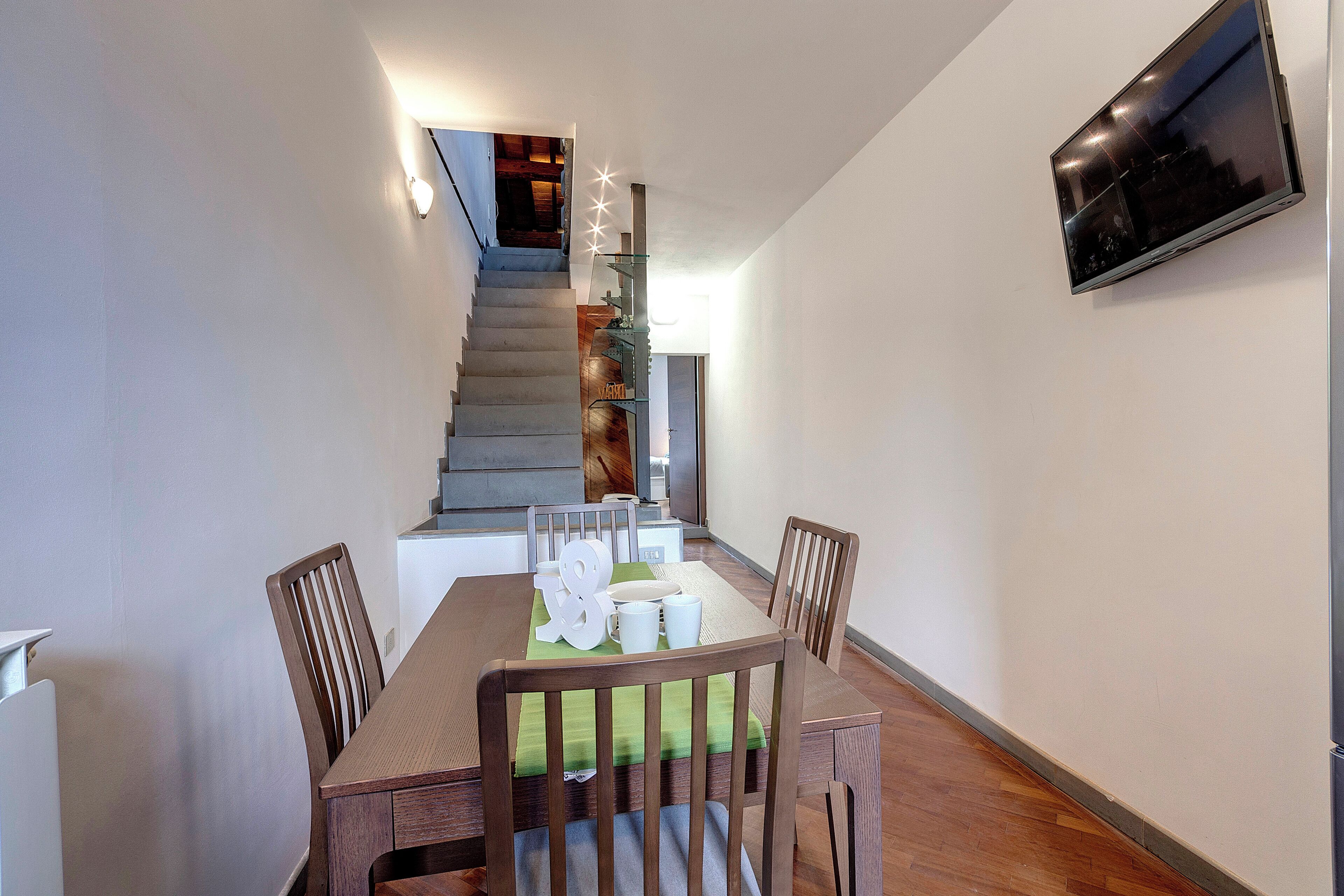 Un comedor con una mesa de madera, sillas y una escalera al fondo.