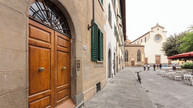 A stone-paved street with a large wooden door and green shutters, leading to a building with a church in the background. - Mamo Florence - Spirito19 Apartment (Florence)