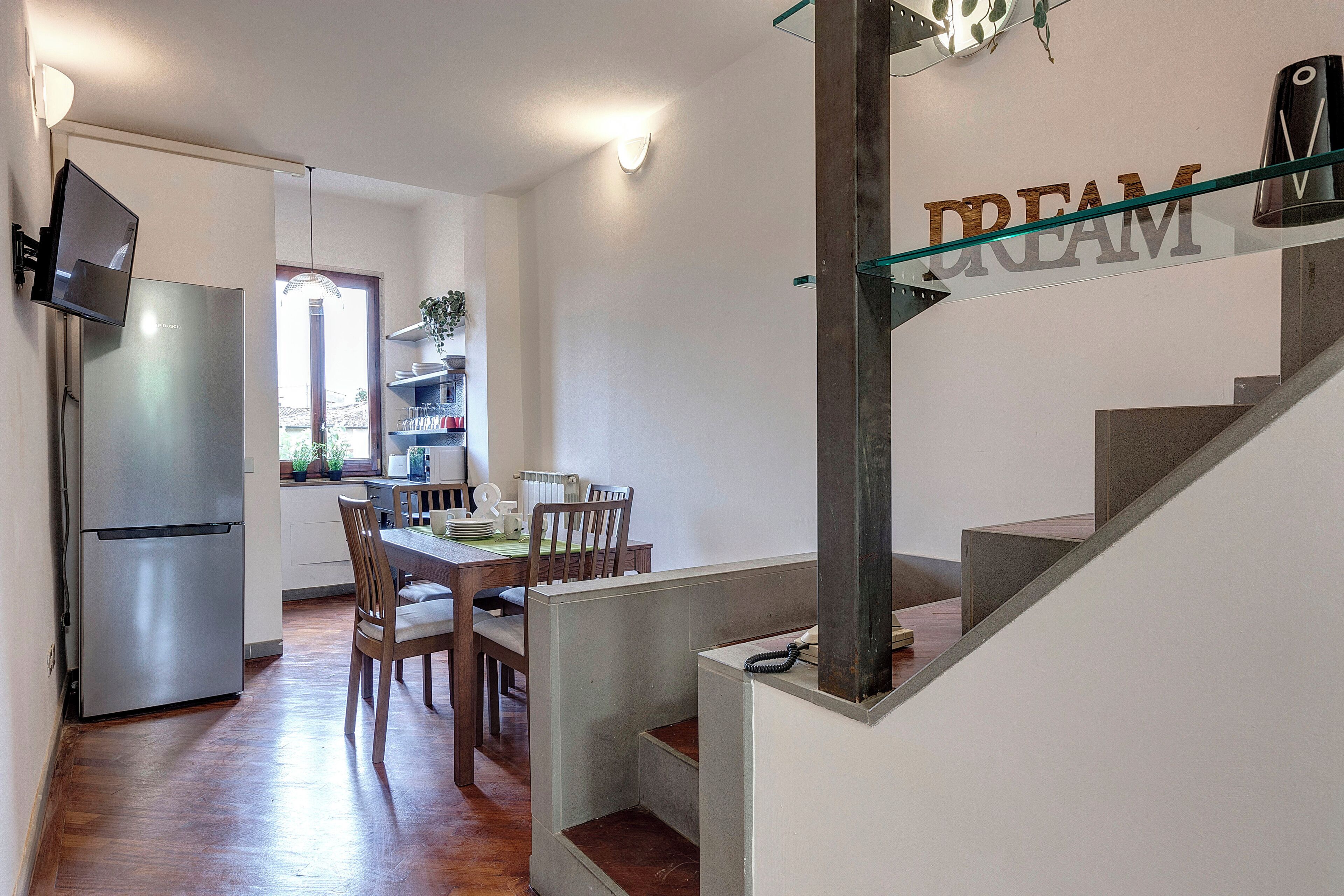 Eine moderne Küche mit einem Edelstahlkühlschrank, einem Holztisch und Stühlen. Eine Treppe mit Glastrennwand führt zu einer Plattform mit dem Schild 'DREAM'.