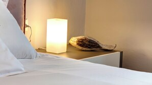 Uma cama com lençóis brancos, uma mesa de cabeceira com um abajur retangular e um objeto decorativo sobre a mesa.