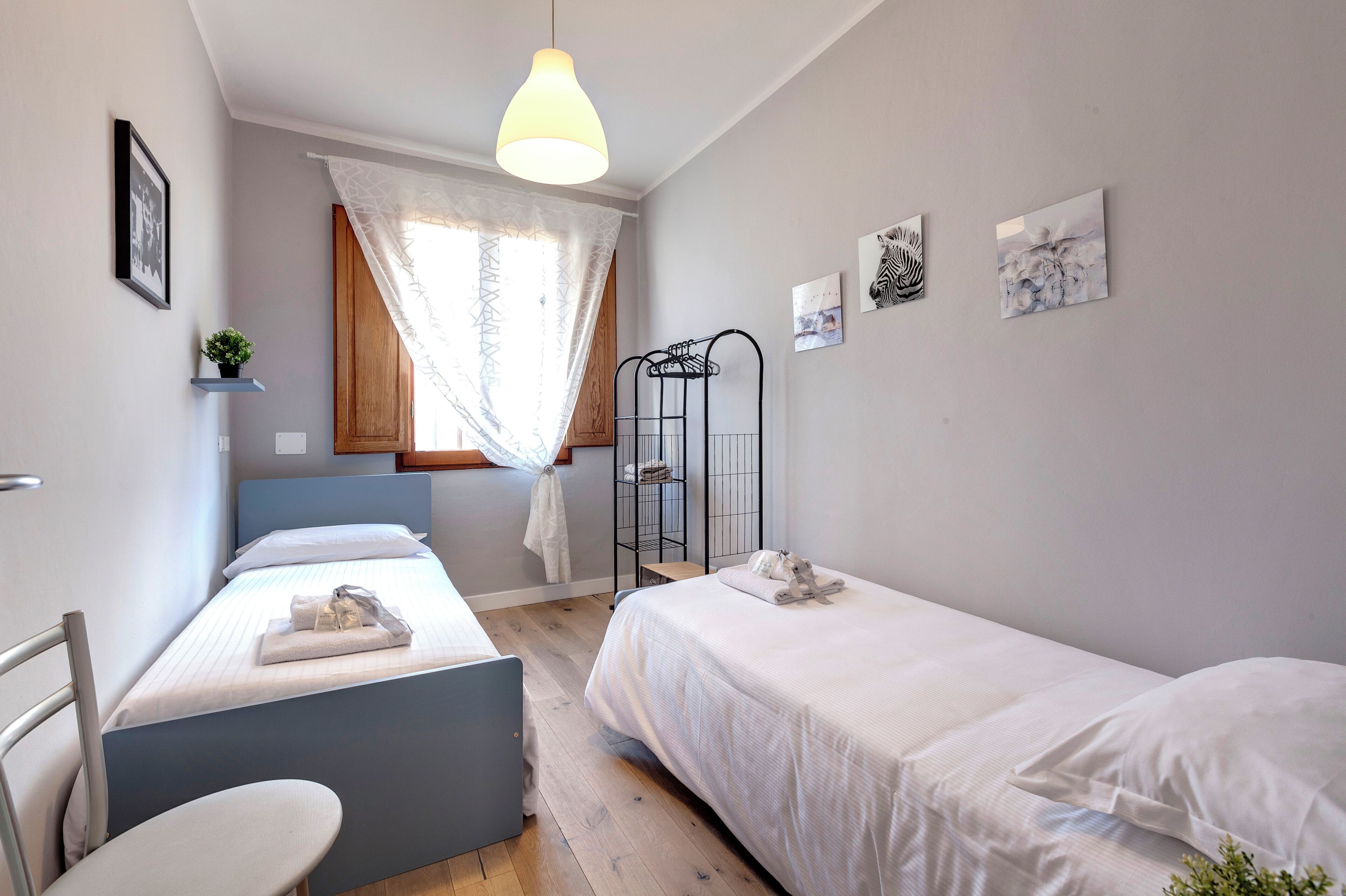 A bedroom with two beds, a desk, a chair, a hanging light, a window with curtains, and wall art.