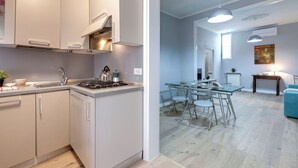 Cozinha moderna com armários brancos e fogão a gás, sala de jantar com mesa de vidro e cadeiras de metal, e sala de estar com sofá azul e mesa de centro de madeira.