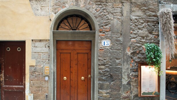 Un edificio de piedra con puerta de madera, una pequeña placa con el número 15, y dos macetas con plantas en el suelo.