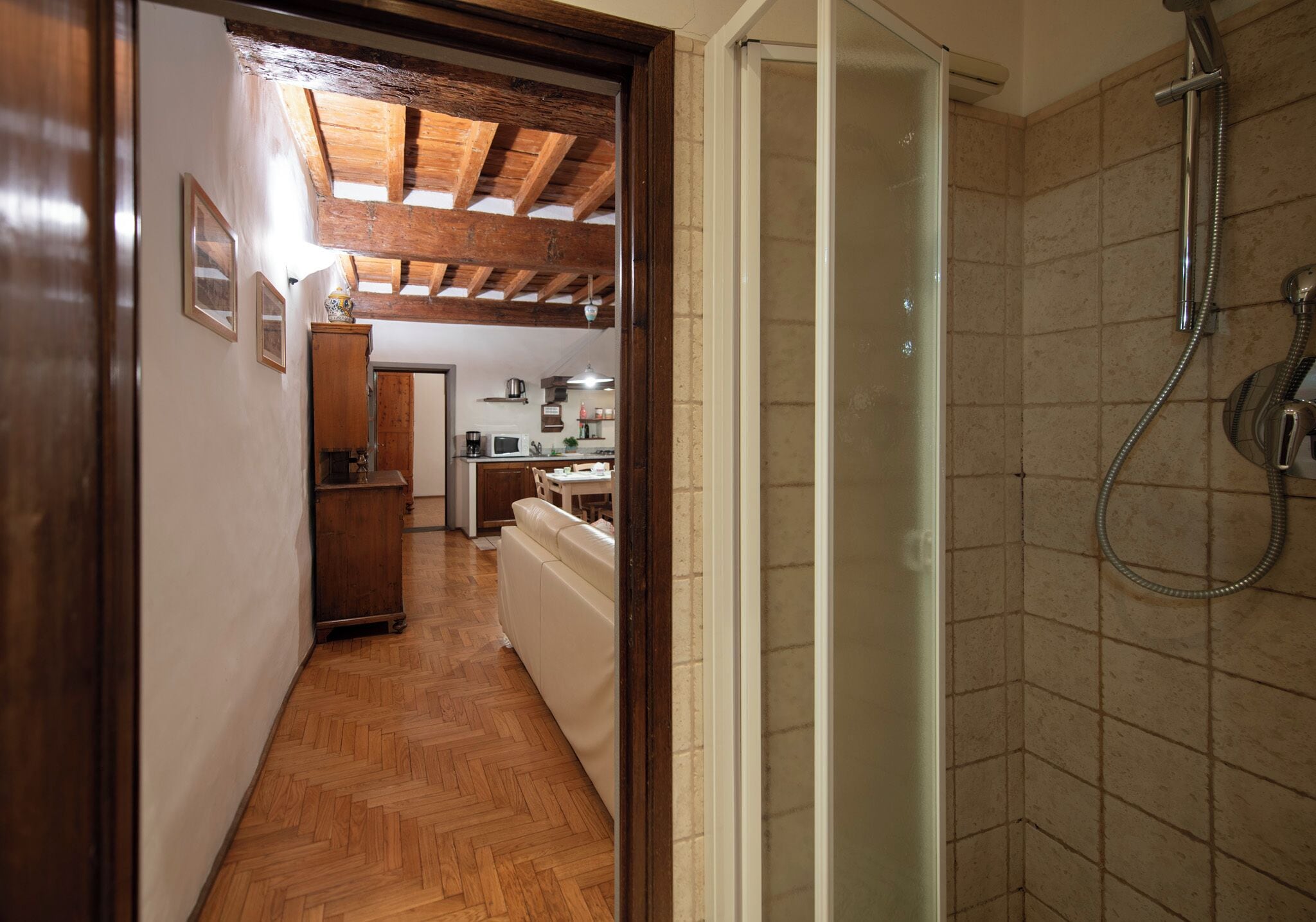 A hallway leading to a bathroom with a shower, wooden flooring, and a kitchen area visible in the background.