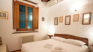 Una cama bien hecha con sábanas y toallas blancas, cabecera de madera, ventana con contraventanas de madera y varias fotografías enmarcadas en la pared.