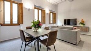 A modern living room with a dining area, featuring a sofa, TV, and a dining table with chairs. - Mamo Florence - Venere & Zefiro Apartments (Florence)