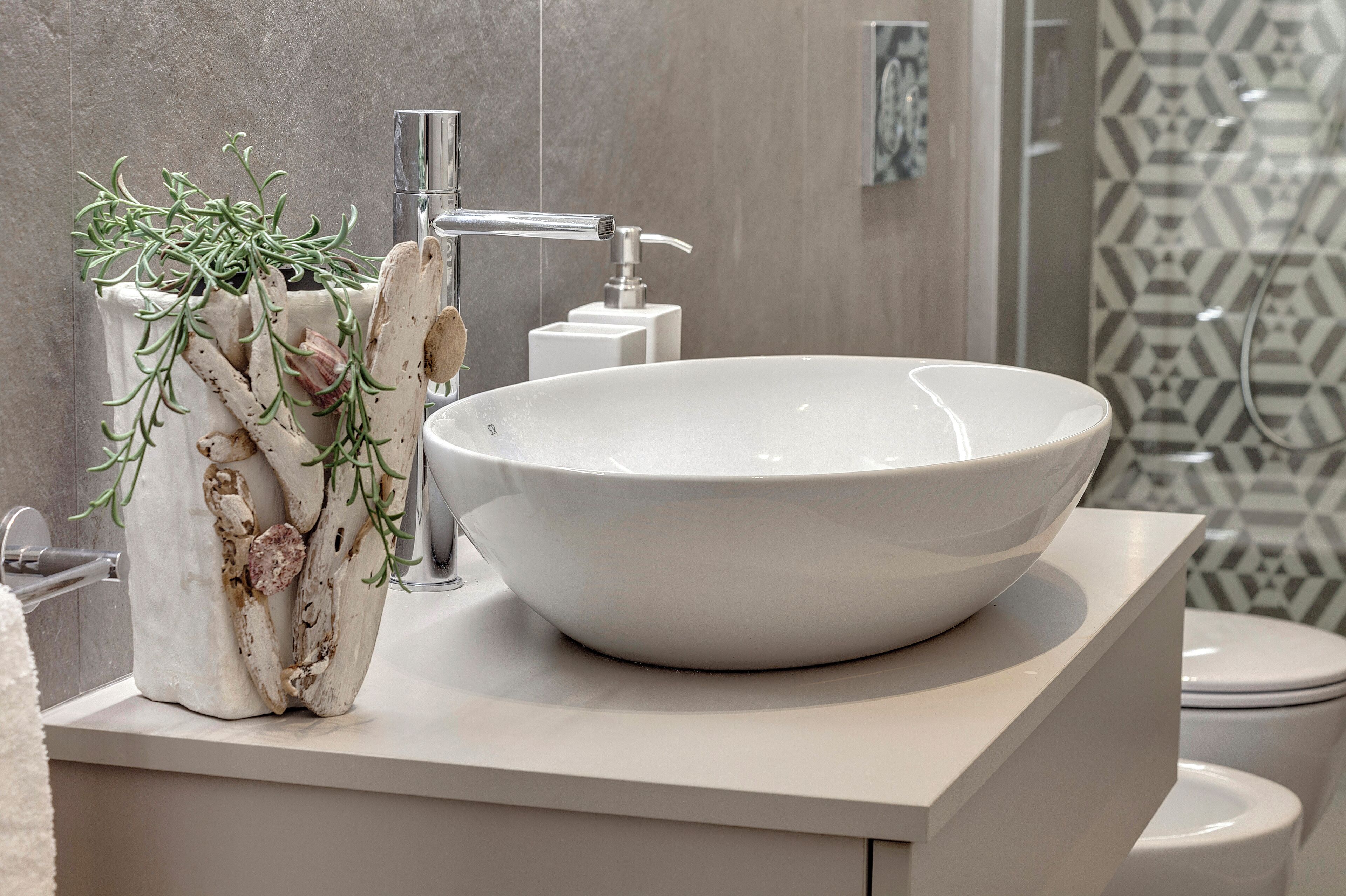 A modern bathroom with a large white vessel sink, a white pedestal with a plant, a chrome faucet, and a toilet.
