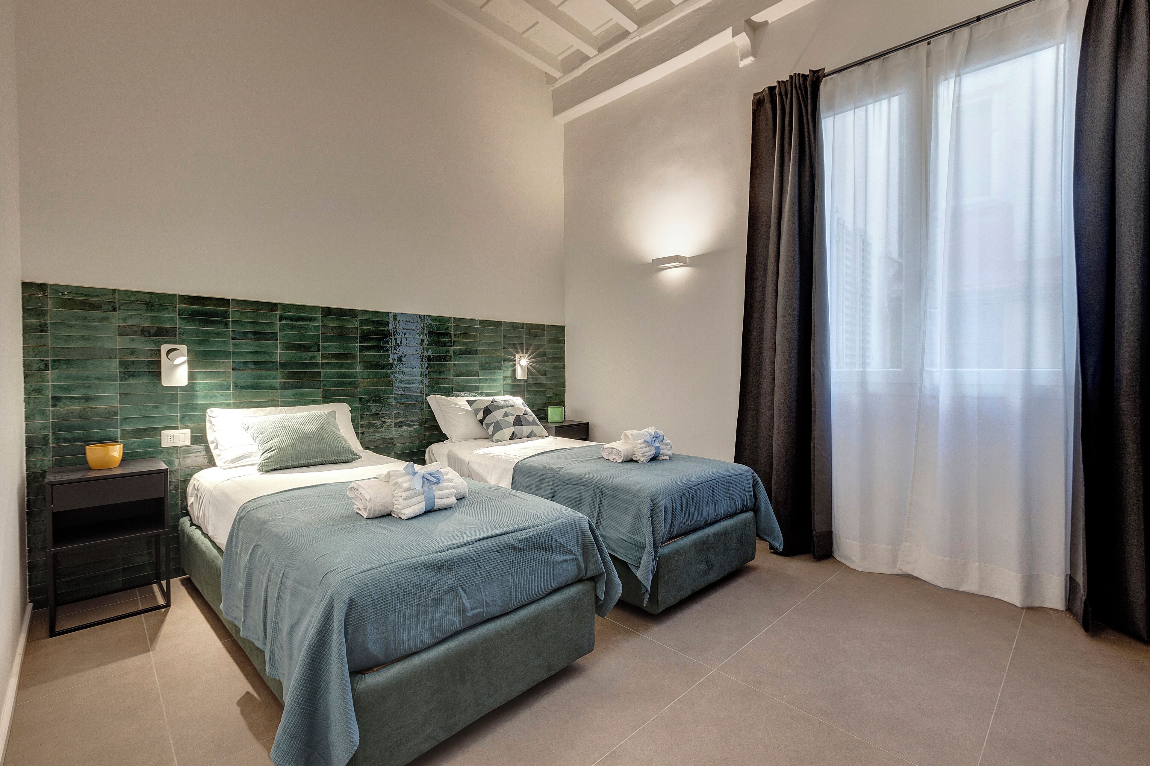 A hotel room with two beds, a green-tiled headboard, a nightstand with a bowl, and a large window with curtains.