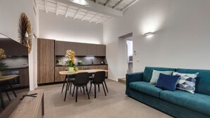 A modern living room with a teal sofa, a dining area with a wooden table and chairs, and a kitchen with wooden cabinets and a white ceiling. - Mamo Florence - Venere & Zefiro Apartments (Florence)