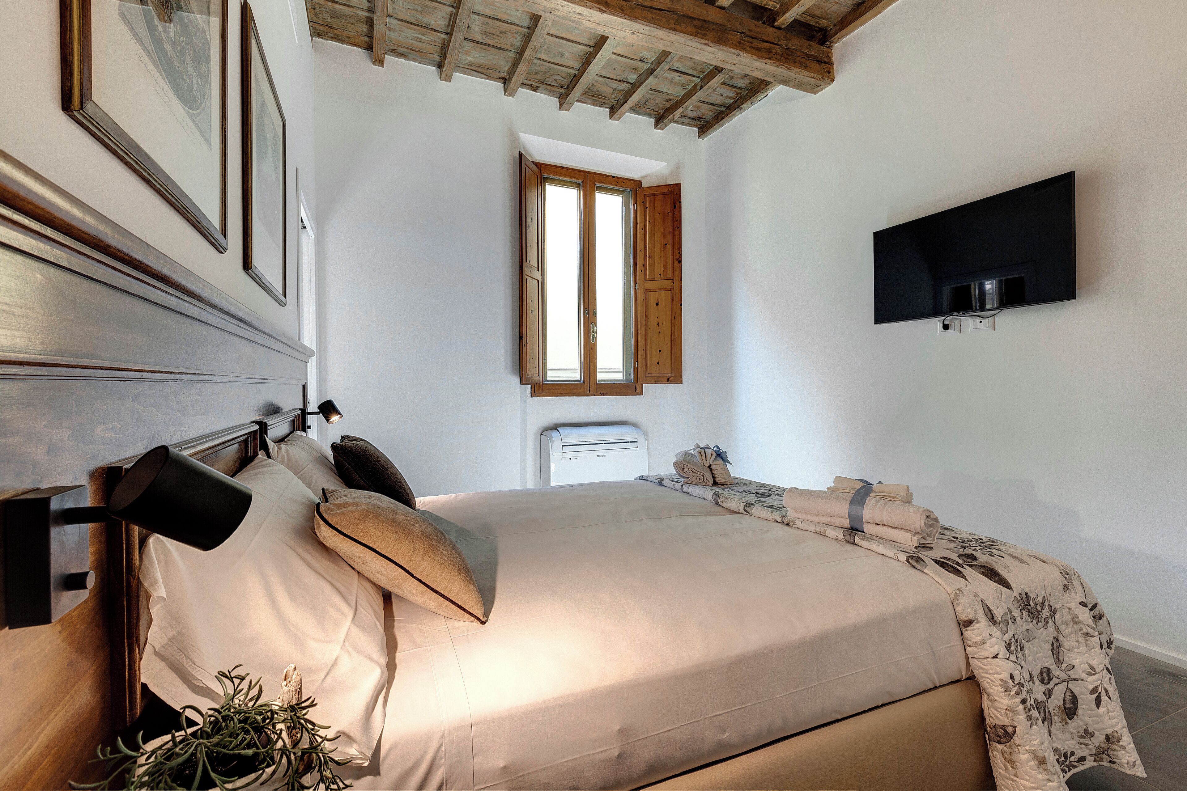 A bedroom with a bed, a mounted TV, a window, and a ceiling with exposed beams.