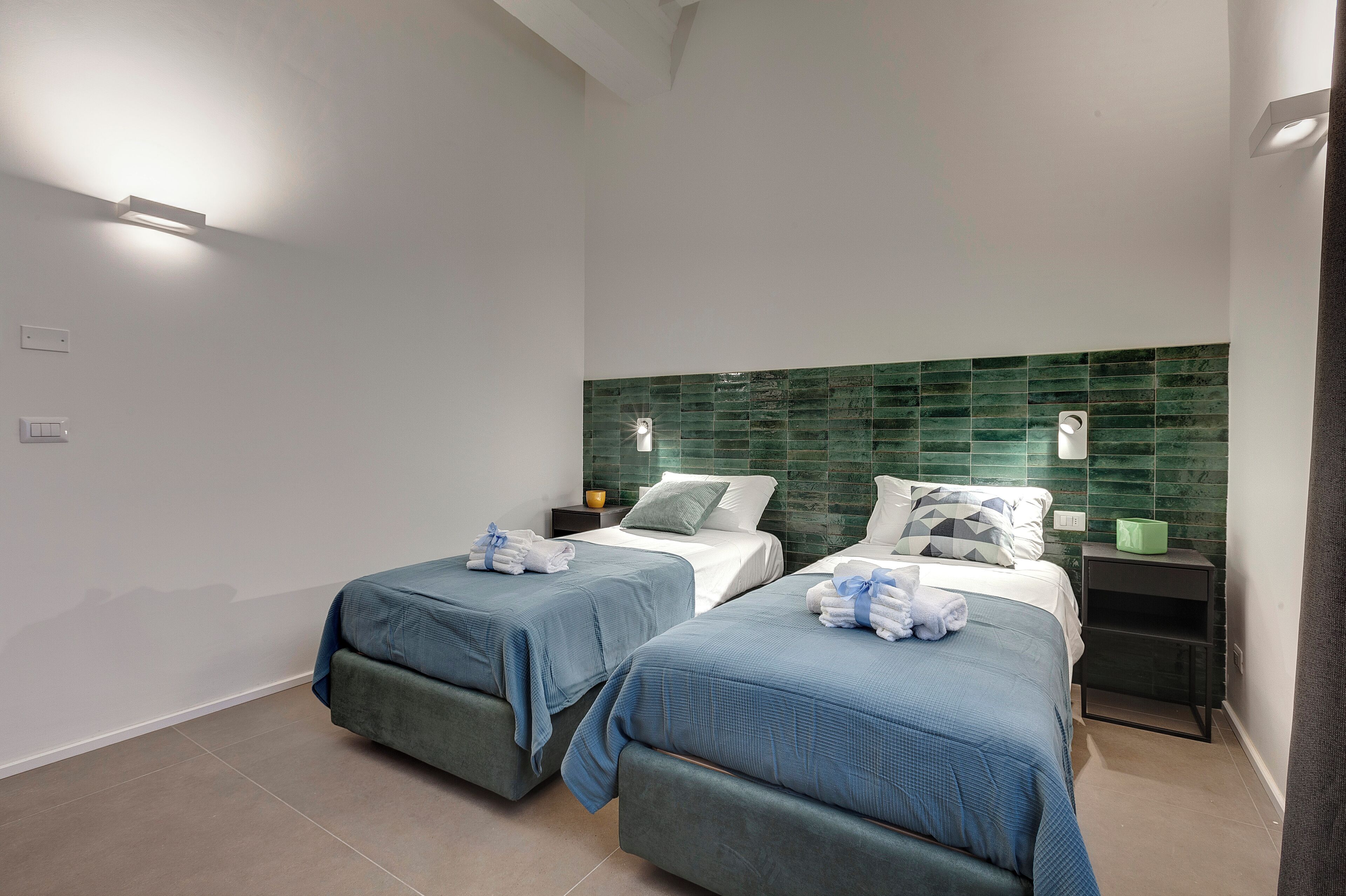 A hotel room with two beds, a green tiled headboard, bedside tables with lamps, and a tiled floor.