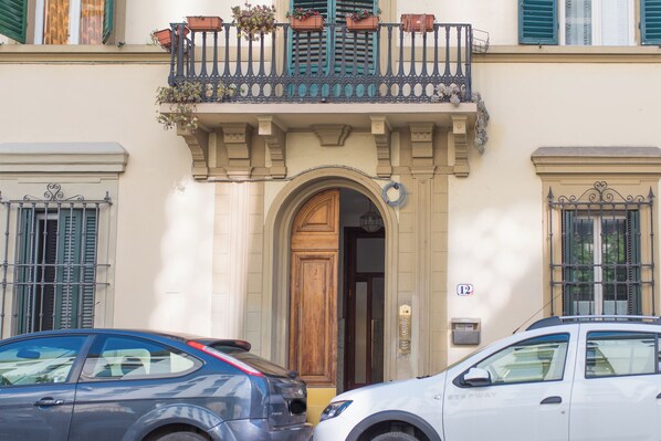 A building with a wooden door, a balcony with flower boxes, and two cars parked in front. - Mamo Florence - Cittadella Apartment (Florence)