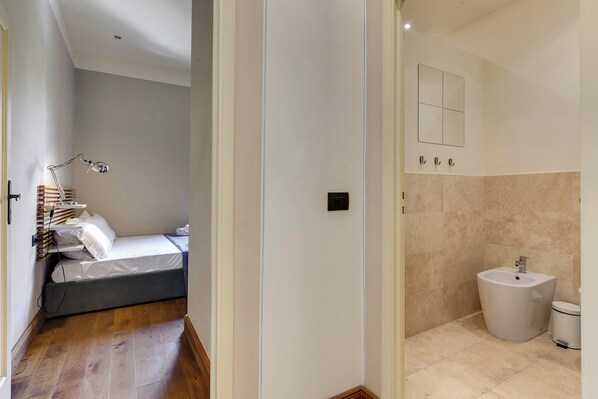 A bedroom with a bed, bedside table, lamp, and a small desk with books. The bathroom has a sink, mirror, and toilet.