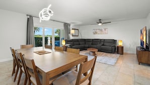 A dining area with a wooden table and chairs, a sofa, a ceiling fan, and a TV on a wooden stand. - Fifty Gulfside Condominiums (Indian Rocks Beach)