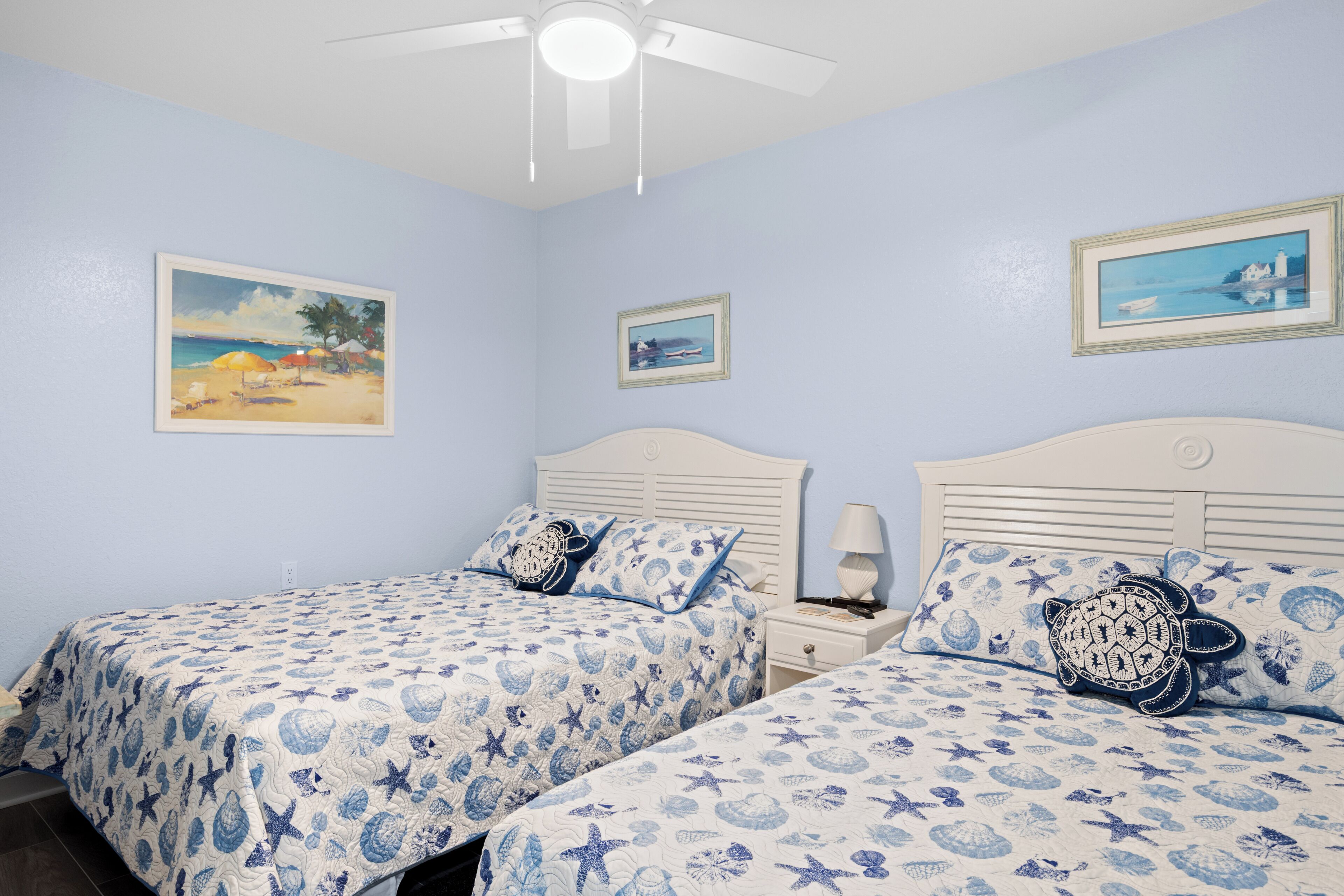 A bedroom with two beds, a ceiling fan, and wall art depicting beach scenes.