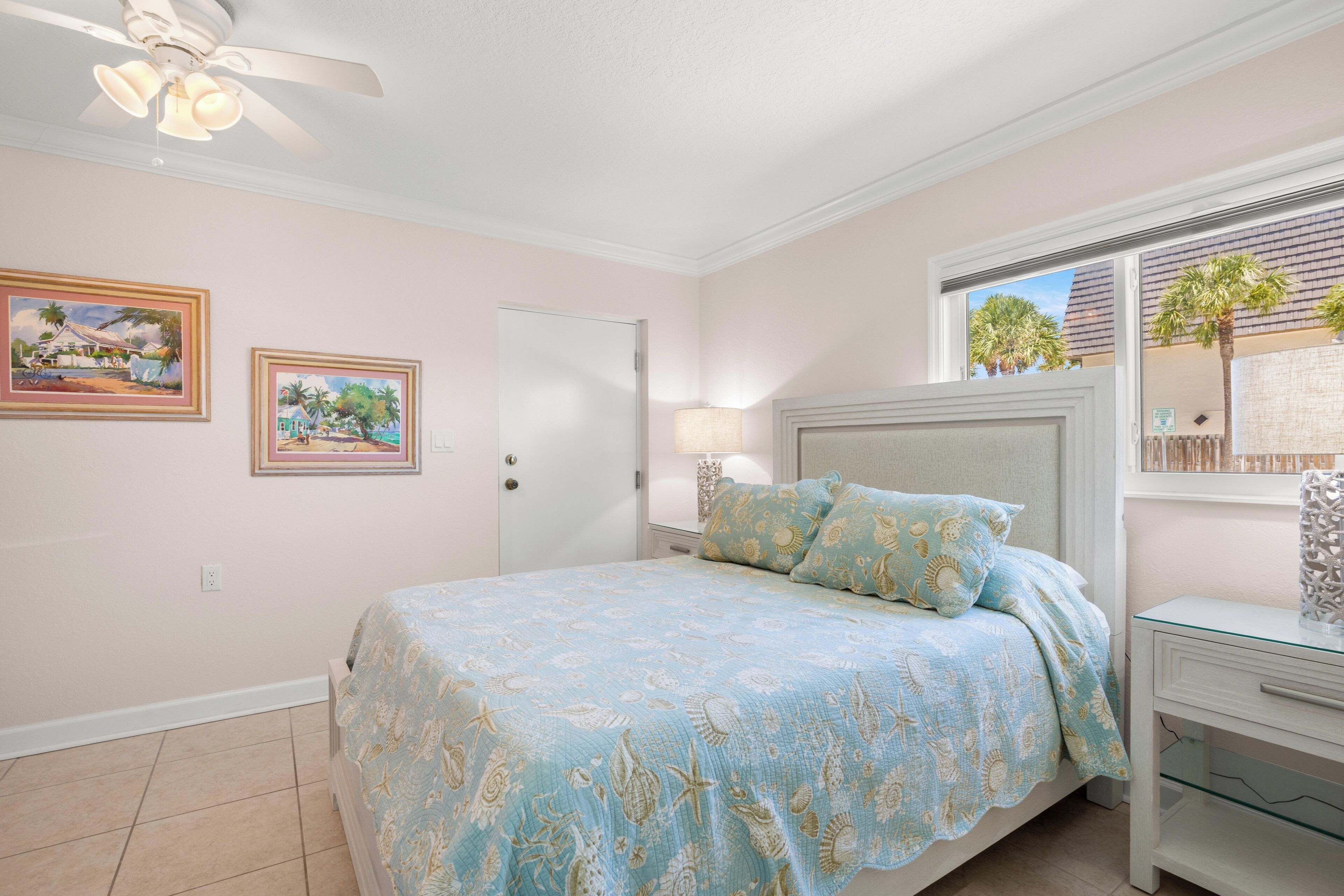 A bedroom with a bed, a nightstand, a ceiling fan, a door, and two framed pictures on the wall.