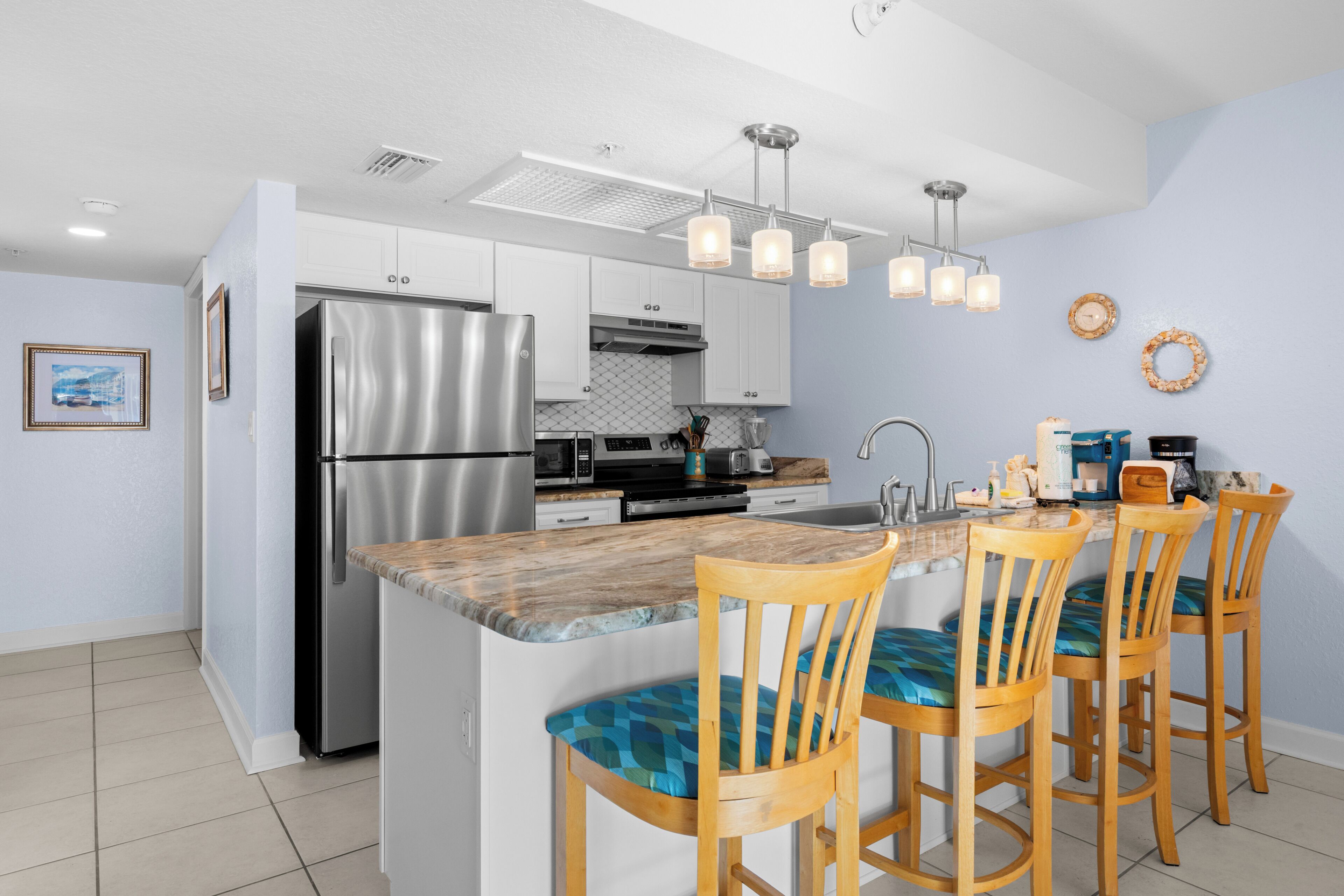 A modern kitchen with a stainless steel refrigerator, a built-in oven, a microwave, and a coffee maker. There are wooden chairs with blue cushions around a central island.