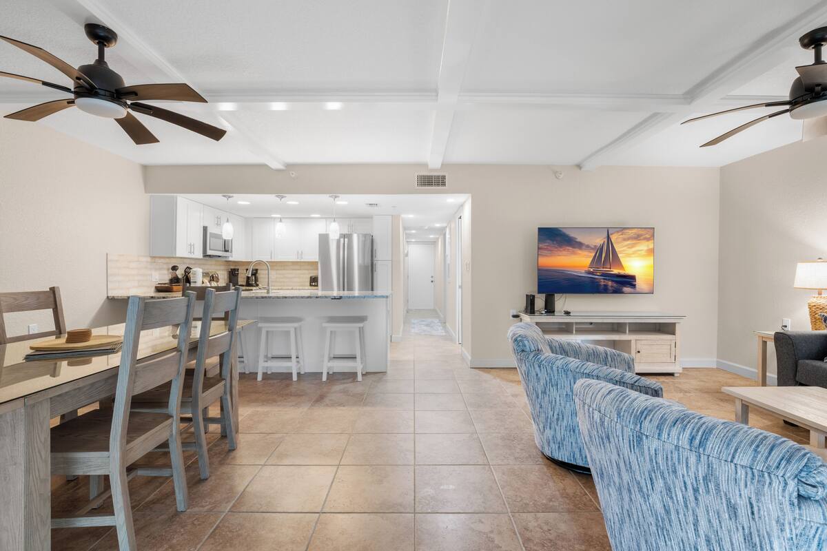 Une cuisine moderne avec un îlot central, un coin repas avec des chaises en bois, un canapé bleu à motifs et un téléviseur diffusant une scène de coucher de soleil.