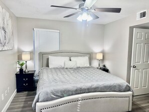 A bedroom with a bed, two bedside tables, a ceiling fan, a window with blinds, and a door. - I040 - Cozy 1st Floor 2BD | DFW South | Irving (Irving)
