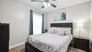 A bedroom with a bed, a nightstand, a ceiling fan, a window with curtains, and a painting on the wall. - I040 - Cozy 1st Floor 2BD | DFW South | Irving (Irving)