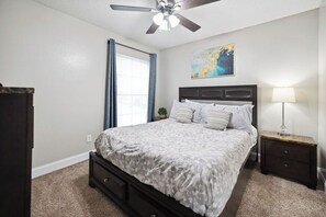 Une chambre à coucher avec un lit, une table de chevet, un ventilateur de plafond, une fenêtre avec des rideaux et un tableau accroché au mur.