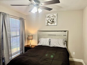 A bedroom with a bed, a ceiling fan, a window with curtains, a bedside lamp, and a wall-mounted artwork.