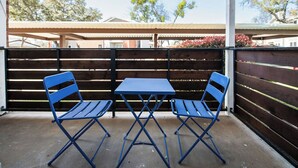 Eine Terrasse mit zwei blauen Klapptischen und Stühlen, einem Holzzaun und einem überdachten Bereich im Hintergrund.