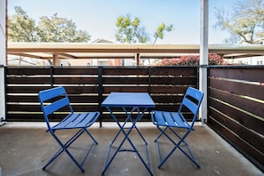 A patio with two blue folding tables and chairs, a wooden fence, and a covered area in the background. - I040 - Cozy 1st Floor 2BD | DFW South | Irving (Irving)