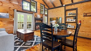 A wooden interior with a fireplace, dining table, chairs, and a ceiling fan. - Couples Cabin | Screened Hot Tub, Outdoor TV & King Bed · Near Helen, GA (Sautee Nacoochee)