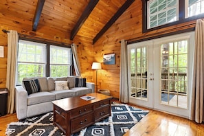 A log cabin interior with a sofa, coffee table, and a large window with a view of greenery. - Romantic Couples Cabin: Screened Hot Tub + Fireplace + Forest Views - Near Helen (Sautee Nacoochee)