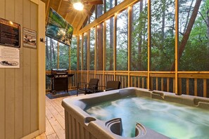 A hot tub in a wooden deck with a view of a forest, a grill, and a chair. - Couples Cabin | Screened Hot Tub, Outdoor TV & King Bed · Near Helen, GA (Sautee Nacoochee)