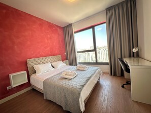 A hotel room with a bed, a desk, and a view of buildings through a window. - MILO Lux Apartments 2 - Belgrade
Waterfront - Aqua (Beograd)