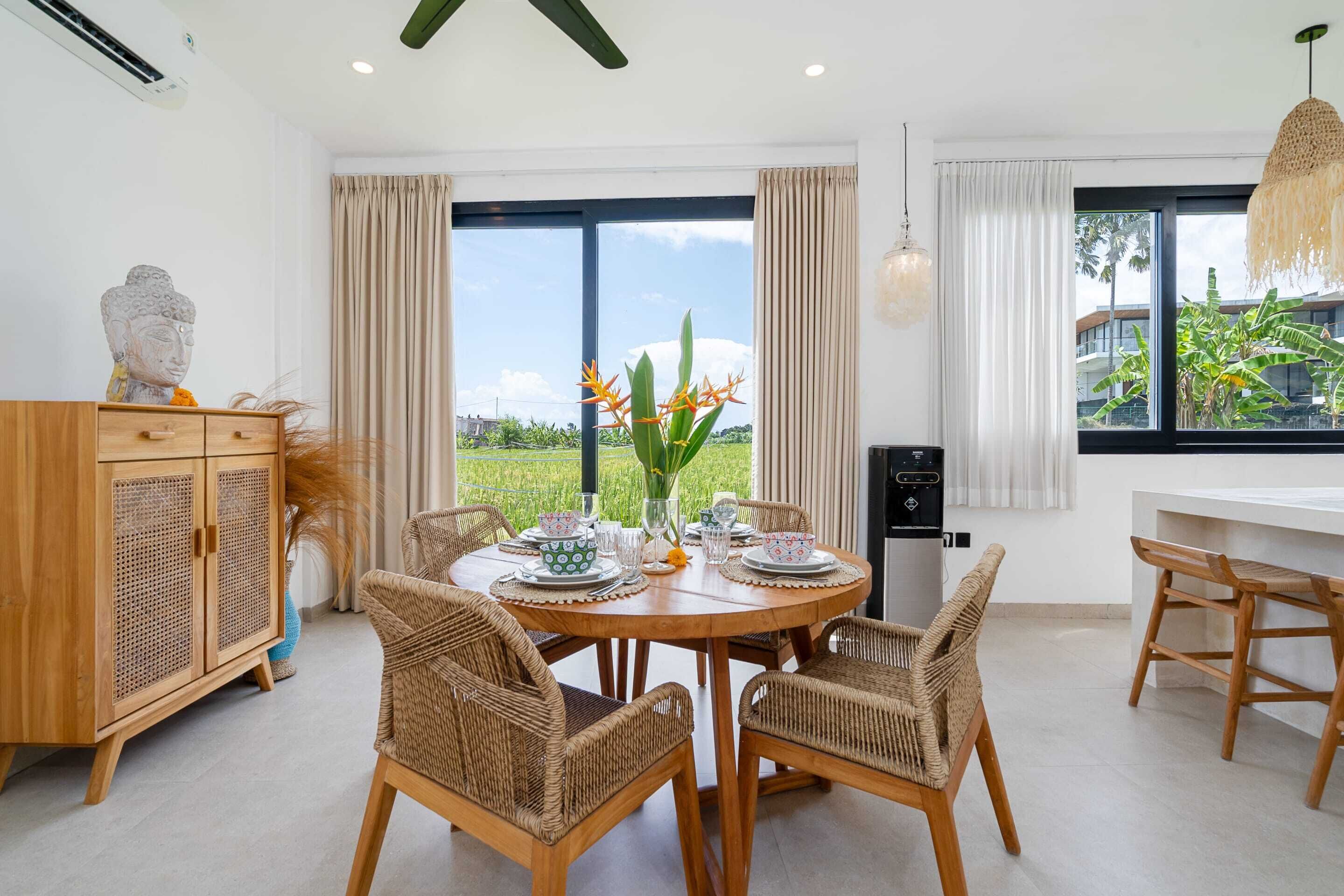Un coin repas avec une table ronde en bois, des chaises en osier, un meuble orné d’une statuette de Bouddha et une vue sur la verdure environnante.