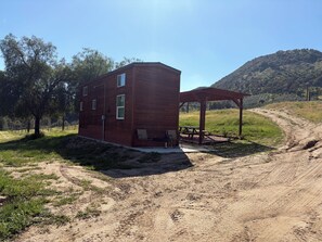 A rustic cabin with a wooden exterior, a covered patio with a table and chairs, and a dirt path leading to it. - Malbec Cabin @ Vinyard Winery, 3 Adults, 1 Kid, WiFi, AC, Fallbrook (Fallbrook)