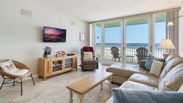 Ein Wohnzimmer mit einer Couch, einem Holztisch, einem Korbstuhl, einem Schrank und einem Fernseher. Ein großes Fenster bietet Blick auf den Strand.
