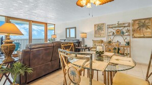 Una sala de estar con un sofá de cuero marrón, una mesa de comedor de vidrio con sillas, un ventilador de techo y vistas al cielo a través de amplios ventanales.