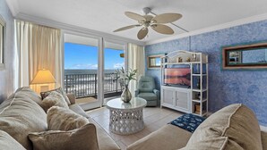 Uma sala de estar com sofá, mesa de centro redonda, ventilador de teto, poltrona, aparador para TV e vista para o mar através de uma porta deslizante.