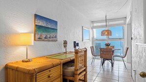 Uma sala de jantar com uma mesa de madeira, cadeiras, uma luminária e um quadro com uma cena de praia.