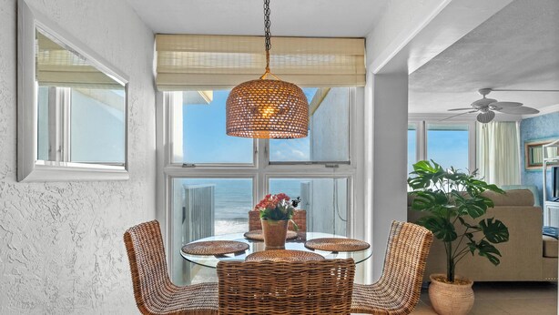 Uma área de refeições com cadeiras de vime e uma mesa de vidro, uma luminária trançada pendente e vista para o oceano através de grandes janelas.