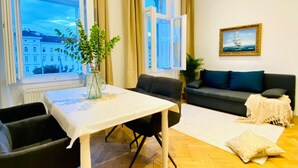 Una sala de estar con una mesa, sillas, un sofá, una ventana con cortinas, una planta y un cuadro en la pared.