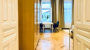 Una habitación con piso de madera en patrón de espiga, una mesa blanca con dos sillas y vistas a través de una ventana con cortinas.