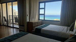 Une chambre d’hôtel dotée d’une grande fenêtre donnant sur la mer, d’un lit, d’une commode et d’une porte-fenêtre coulissante qui mène à un balcon équipé de chaises.