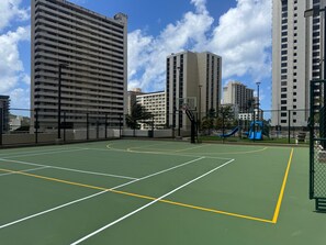 An outdoor basketball court with a blue slide and a basketball hoop, surrounded by tall buildings.