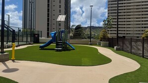Une aire de jeux avec un toboggan et une structure d’escalade, entourée d’une pelouse bien entretenue et avec de grands immeubles en arrière-plan.