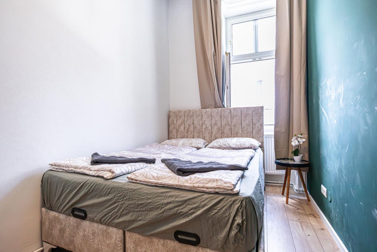 Ein Schlafzimmer mit einem Bett, einem Fenster mit Vorhängen, einem kleinen Tisch mit Pflanze und einer grünen Wand.